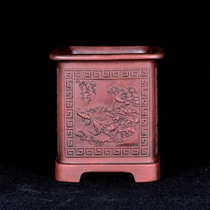 May include: A reddish-brown, rectangular planter with a detailed relief carving of a landscape scene. The planter features a square top and a decorative border. The carved scene includes fish, plants, and a tree.