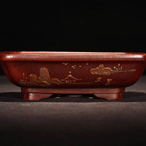 May include: A rectangular red bonsai pot with a gold painted landscape design. The pot has four small feet.