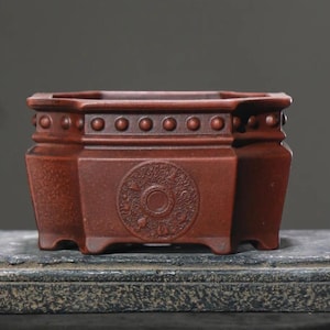 May include: A reddish-brown, hexagonal ceramic planter with a detailed, embossed design. The planter features a series of small, raised circles along the top edge and a circular, ornate pattern on the front. The overall design is symmetrical and detailed.