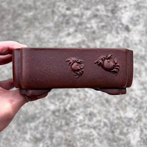 May include: A rectangular, reddish-brown ceramic bonsai pot. The pot features two crab designs on the front. The pot has a raised base and is held by a hand against a blurred background.