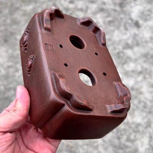 May include: A reddish-brown, square bonsai pot with a textured surface. The pot has a rectangular shape with rounded edges and decorative relief carvings. It features drainage holes and is designed for planting small trees.