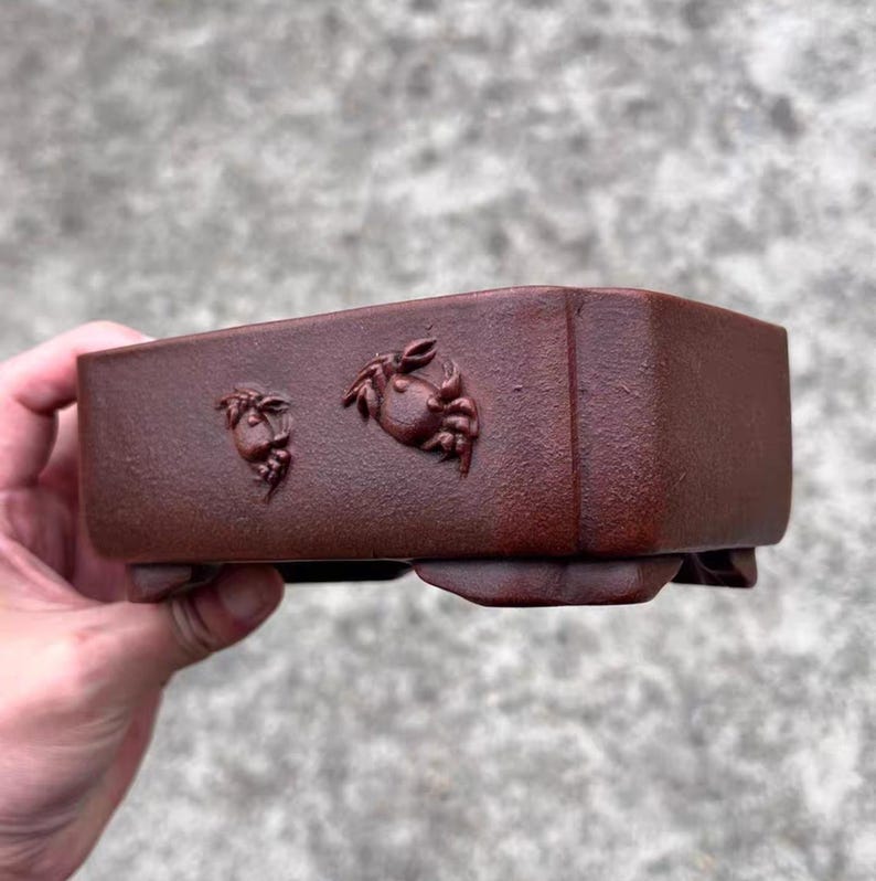 May include: A rectangular, reddish-brown bonsai pot with crab relief carvings. The pot has a textured surface and is supported by small feet. The crab carvings are on the front of the pot.