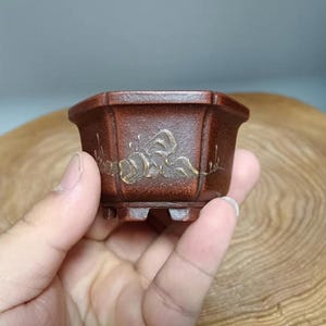 Puede incluir: Una pequeña maceta de bonsái octogonal de terracota con un esmalte marrón y detalles en pan de oro. La maceta tiene cuatro pequeñas patas.