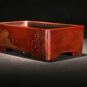 May include: A rectangular, dark red bonsai pot with a raised lip and four small feet. The pot has a decorative design of stylized clouds and mountains in gold.