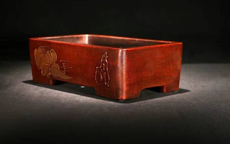 May include: A rectangular, dark red bonsai pot with a raised lip and four small feet. The pot has a decorative design of stylized clouds and mountains in gold.