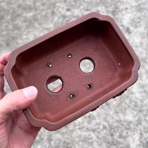 May include: A rectangular, reddish-brown bonsai pot with rounded edges and drainage holes. The pot is made of a terracotta-like material and has a slightly textured surface. The pot is held by a person's hand.