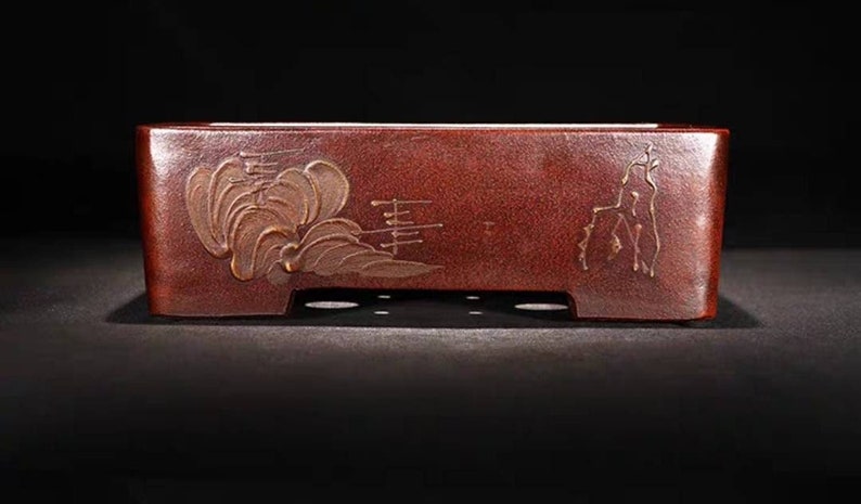 May include: A rectangular, dark red ceramic bonsai pot with a raised, gold-colored design of stylized clouds and a few vertical lines. The pot has drainage holes on the bottom.
