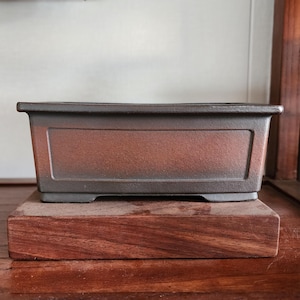 Könnte beinhalten: Ein rechteckiger Bonsai-Topf mit dunkelbrauner, strukturierter Oberfläche. Der Topf steht auf einem Holzsockel mit rötlich-braunem Farbton. Das Design des Topfes weist eine leicht erhöhte, rechteckige Platte auf der Vorderseite auf.