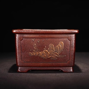 May include: A rectangular, dark red ceramic planter with a gold-colored landscape design. The planter has a square base and a slightly raised rim.