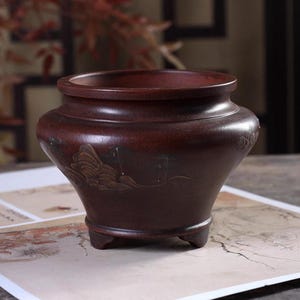 May include: A dark brown, matte-finished ceramic vessel with a wide, rounded body, a flared rim, and three small feet. The exterior surface features a subtle, engraved landscape design depicting mountains and trees in a monochromatic style.