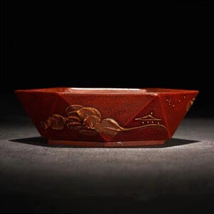 Pode incluir: Um vaso octogonal de cerâmica vermelha com um design de paisagem pintado a ouro. O vaso tem uma superfície texturizada e uma borda ligeiramente alargada.