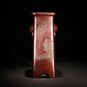 Puede incluir: Un jarrón alto, rectangular, de cerámica de color marrón rojizo con diseños dorados. El jarrón tiene una base cuadrada y dos asas en forma de cabeza de león a los lados. El diseño presenta nubes estilizadas y una inscripción vertical en oro.