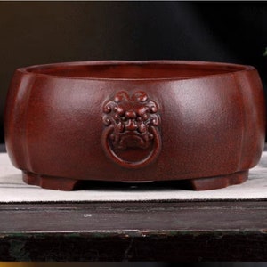 Puede incluir: Una maceta redonda de color terracota con un diseño de cabeza de león en la parte delantera. La maceta tiene tres patas y está hecha de un material texturizado.