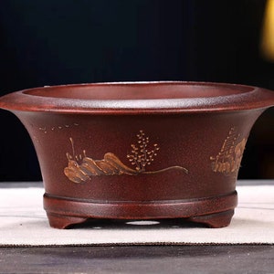 May include: A dark red ceramic bonsai pot with a gold painted landscape design. The pot has three small feet.