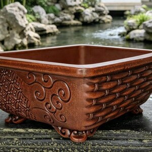 May include: A square, brown ceramic planter with decorative detailing. The planter has a woven basket design on the sides and ornate patterns on the corners. It stands on small, curved feet. The background shows a tranquil garden setting.