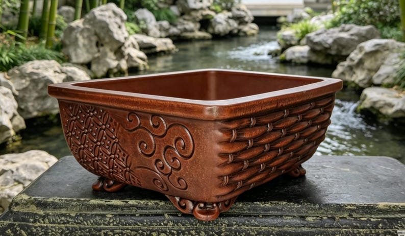 May include: A square, brown ceramic planter with decorative detailing. The planter has a woven basket design on the sides and ornate patterns on the corners. It stands on small, curved feet. The background shows a tranquil garden setting.