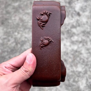 May include: A rectangular, dark brown ceramic object with two crab relief carvings. The object has a textured surface and is held by a person's hand. The crabs are detailed with claws and eyes.