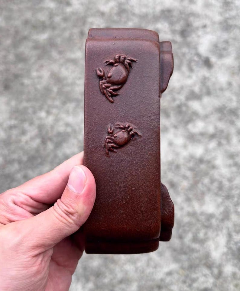 May include: A rectangular, dark brown ceramic object with two crab relief carvings. The object has a textured surface and is held by a person's hand. The crabs are detailed with claws and eyes.