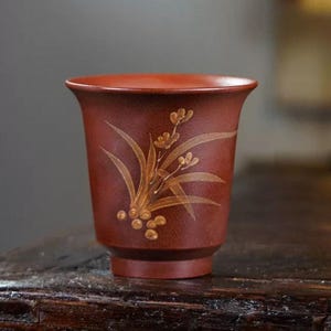 Peut inclure: Une tasse à thé en céramique de couleur marron avec un bord évasé et un petit pied. La tasse est ornée d'un motif floral doré. La tasse est probablement utilisée pour servir du thé ou d'autres boissons chaudes.
