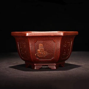 May include: A dark red hexagonal ceramic pot with a textured surface and gold-colored Chinese characters and designs. The pot has a wide base and a slightly flared rim.
