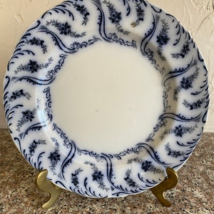 Puede incluir: Un plato de porcelana blanca con un patrón floral azul y blanco. El plato tiene un borde festoneado y está en un soporte dorado.