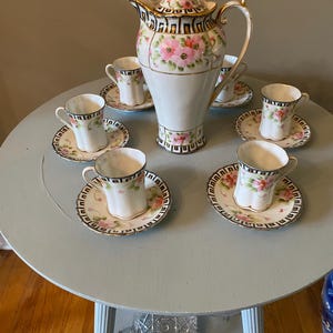 May include: A porcelain tea set consisting of a tall teapot with a lid and five matching teacups on saucers. Each piece features a white base with hand-painted pink flowers, green leaves, and a black and gold geometric Greek key border pattern.