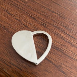 May include: A silver-toned metal heart-shaped object featuring a solid left half and an open, hollow right half. The piece has a smooth, reflective surface and a curved, continuous outer edge that forms the heart silhouette.