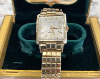 May include: A vintage gold-toned Wittnauer Statesman wristwatch with a rectangular face and a gold expansion band. The watch face is silver with gold hands and markers. The watch is displayed in a green velvet-lined box with gold trim.