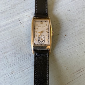Op de afbeelding: Een vintage Bulova polshorloge in goudkleur met een witte wijzerplaat en een zwart leren bandje. Het horloge heeft een rechthoekige wijzerplaat met Romeinse cijfers als uurmarkeringen en een secondewijzer.