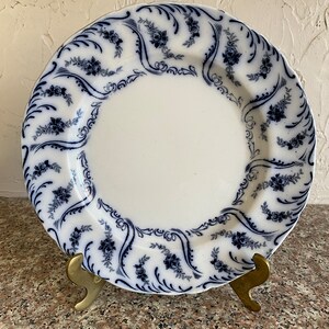 May include: A white ceramic plate with a blue and white floral pattern. The plate has a scalloped edge and is decorated with a repeating design of stylised flowers and leaves.