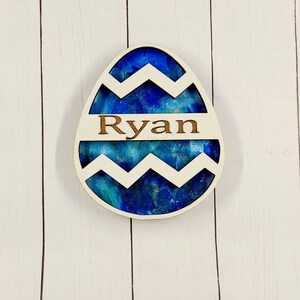 Puede incluir: Un adorno de huevo de Pascua de madera con un diseño marmoleado azul y verde y un patrón de chevron blanco. El nombre "Ryan" está grabado en el huevo.