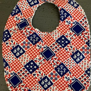 May include: A red, white, and blue baby bib with a patchwork design featuring anchors, sailboats, stars, and gingham. The bib has a ruffled edge.