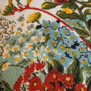 May include: Close-up of a floral textile with a woven texture. The design features a variety of flowers in shades of red, blue, yellow, and green against a white background. The flowers are arranged in a dense, detailed pattern.