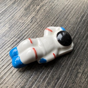 May include: A white ceramic astronaut figurine with red and blue accents. The astronaut is lying on its back with its arms raised above its head.