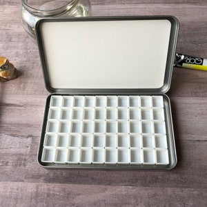 May include: Open metal watercolor paint palette with 48 white plastic wells. The palette is open on a wooden surface, with a watercolor brush, a small owl figurine, and two tubes of watercolor paint.