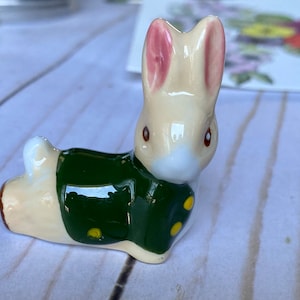 May include: A ceramic bunny figurine, cream-colored with pink inner ears and brown eyes, wearing a green jacket with yellow polka dots. The bunny is lying down, with its head upright. A charming Easter or spring decoration.