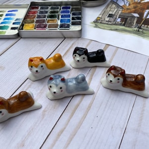 May include: Five ceramic dog-shaped chopsticks rests. The dogs are in various poses and colors, including brown, black, white, and yellow.