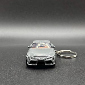 May include: A gray die-cast sports car keychain featuring a detailed front grille, headlights, and a visible interior with a steering wheel. The metal keyring is attached to the side of the vehicle, which displays a small emblem on the hood.