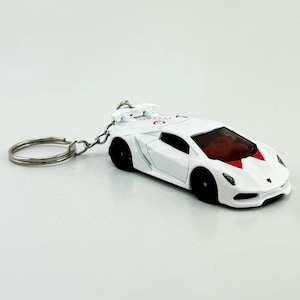 May include: White toy car keychain with black wheels and a red interior. The car has a spoiler and a silver key ring. The car has a logo on the hood.