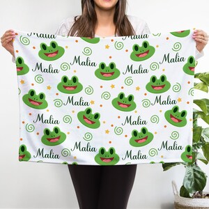 May include: White fleece blanket with a repeating pattern of green frogs, orange swirls, and yellow stars. The name "Malia" is repeated throughout the blanket.