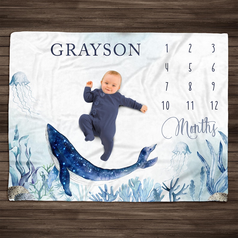 May include: A white baby blanket with a blue whale and coral reef design. The blanket has a numbered grid for tracking a baby's growth. The text "GRAYSON" and "Months" are printed on the blanket. A baby is lying on the blanket.