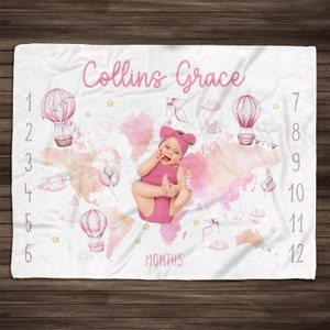 May include: A pink and white baby milestone blanket with a watercolor world map design. The blanket features hot air balloons, stars, and clouds. The text "Collins Grace" and "MONTHS" are printed on the blanket. Numbers 1 through 12 are printed on the right side of the blanket.