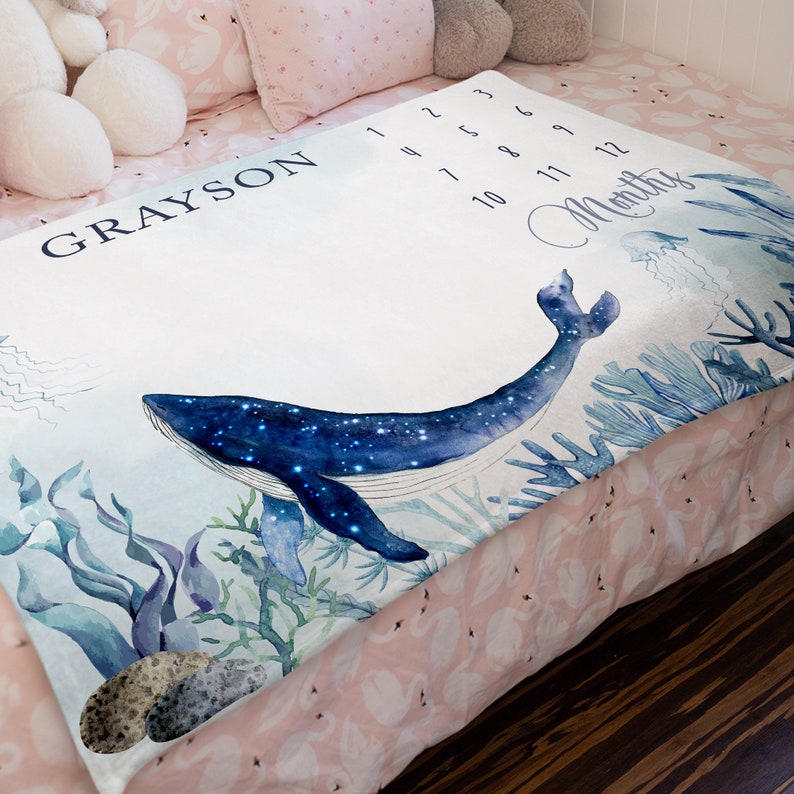 May include: A white baby milestone blanket with a blue whale with stars on it, surrounded by seaweed and coral. The blanket has the name "GRAYSON" and numbers 1-12 for each month. The text "Months" is written in cursive.