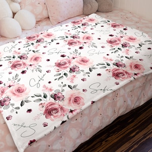 May include: A white fleece blanket with a pink and green floral pattern and the name "Sofia" repeated throughout the design.