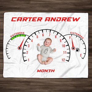 May include: A white baby blanket with a red and black speedometer design. The speedometer is labeled "Cuteness" and has a red needle pointing to the "F" mark. The baby blanket also has the text "CARTER ANDREW" and "MONTH" printed on it.