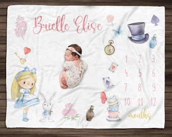 Op de afbeelding: Een wit babydekentje met een Alice in Wonderland-thema. Het heeft een slapende baby, Alice in Wonderland, een wit konijn, een Mad Hatter-hoed, een theekopje en een sleutel. Het dekentje heeft een roze en gouden kleurschema en de tekst "Brielle Elise" en "12 maanden".