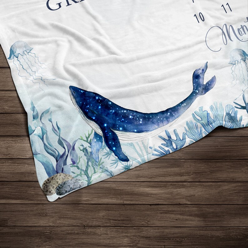 May include: A white baby blanket with a blue whale with stars on it, surrounded by seaweed and coral. The blanket has the numbers 10 and 11 printed on it.