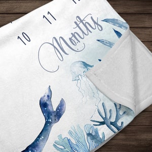 May include: A white baby blanket with a blue watercolor design of a mermaid tail, jellyfish, and coral. The blanket has numbers 10 and 11 printed on it and the text "Months".