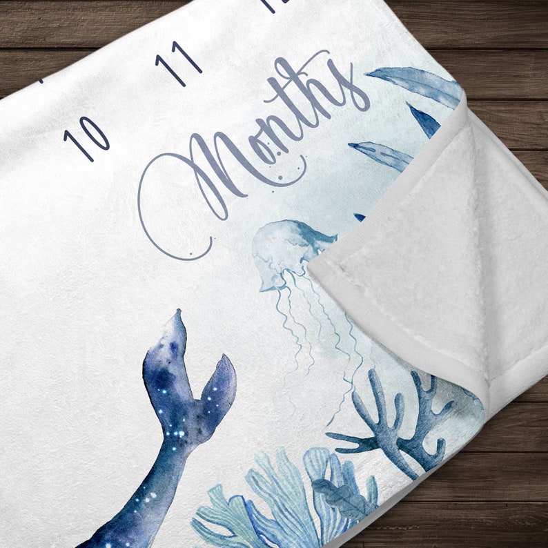 May include: A white baby blanket with a blue watercolor design of a mermaid tail, jellyfish, and coral. The blanket has numbers 10 and 11 printed on it and the text "Months".