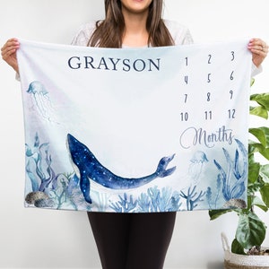May include: A white baby milestone blanket with a blue whale and jellyfish illustration. The blanket has the name "GRAYSON" and numbers 1-12 for tracking monthly growth.